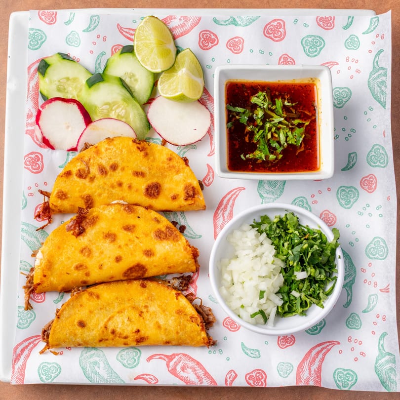 Three golden-fried birria tacos with sides of consome, diced onions, cilantro, cucumbers, radishes, and limes.